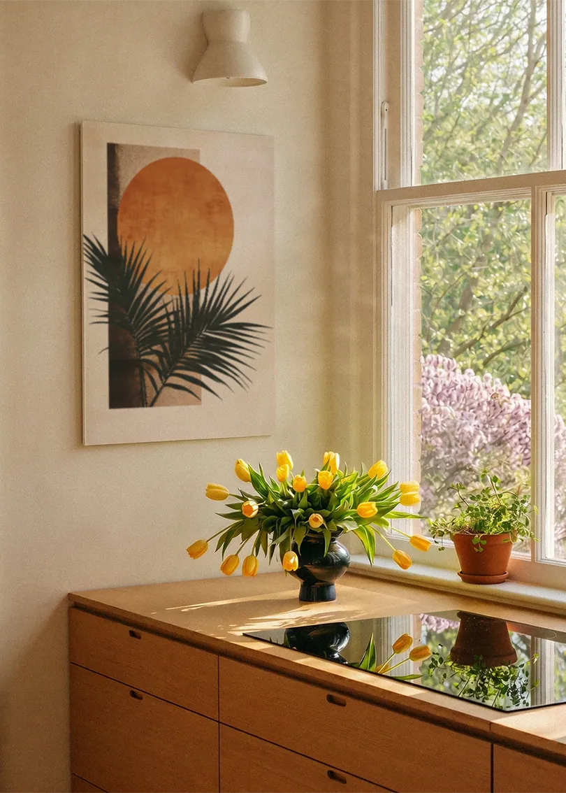 Akustikbillede med en orange cirkel bag sorte palmeblade, ved et vindue med en blomsterbuket i karmen