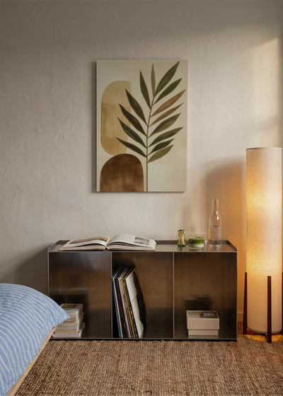 Akustikbillede med et stort grønt blad med beige former, over sengegavlen i et soveværelse med tændt lampe