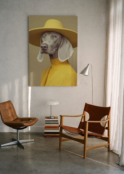 Akustikbillede med en hund med gul hat og gul rullekrave, ophængt over en lænestol ved gardinet med gulvlampe