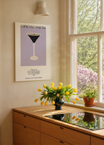 Akustikbillede med en minimalistisk cocktailplakat med Espresso Martini, ved et køkkenvindue med gule tulipaner