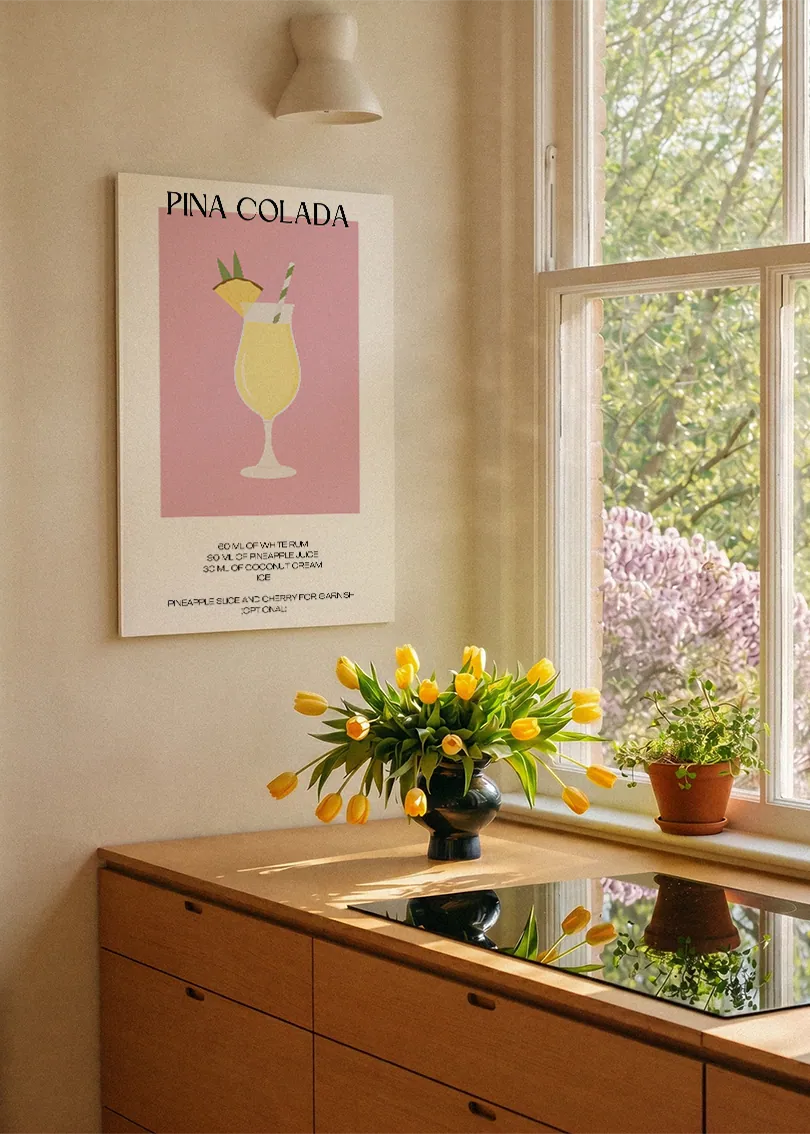 Akustikbillede med en minimalistisk cocktailplakat med Pina Colada, ved et køkkenvindue med gule tulipaner