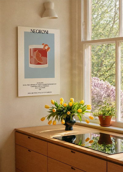 Akustikbillede med en minimalistisk cocktailplakat med Negroni, ved et køkkenvindue med gule tulipaner