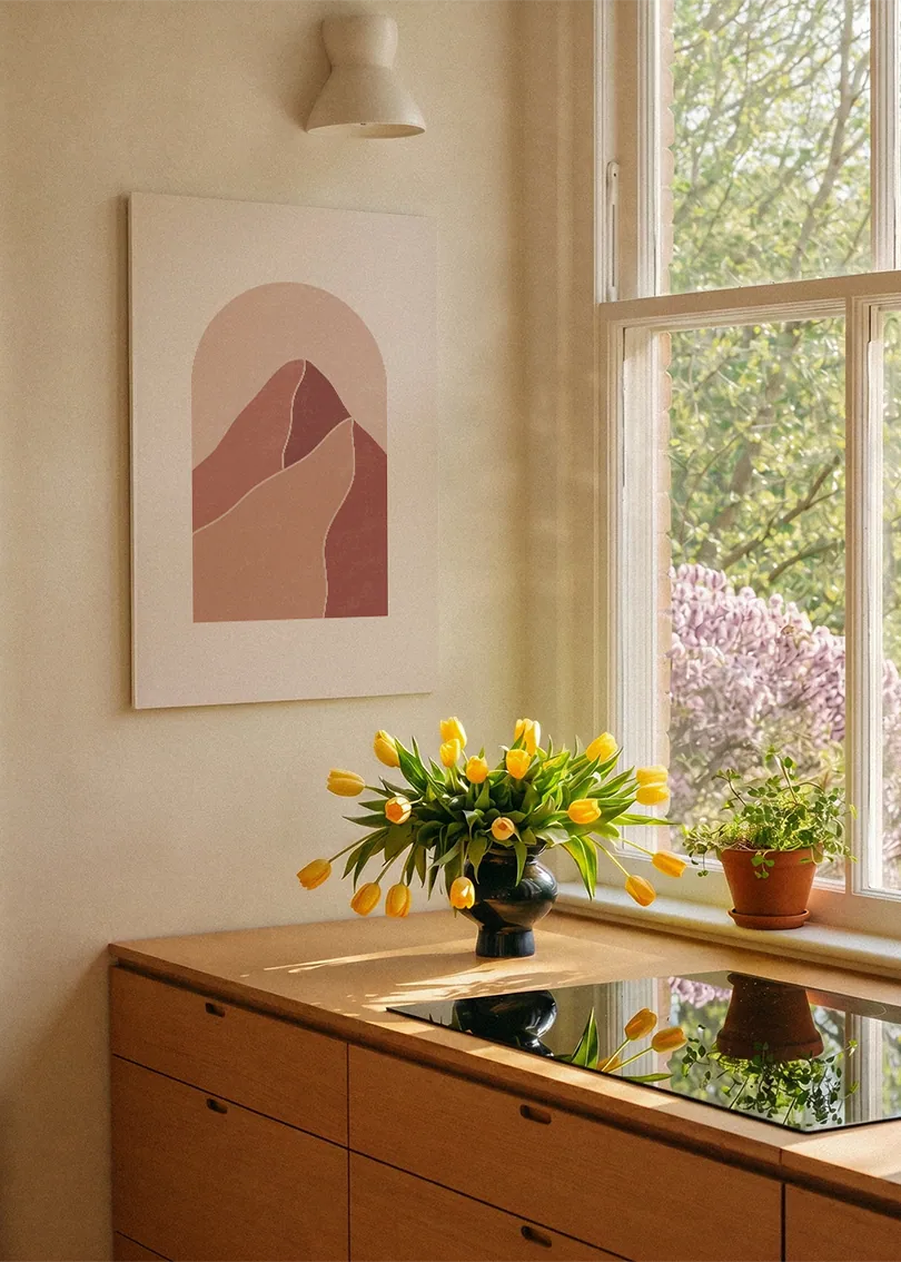Akustikbillede med terrakottafarvede bjerge i en bueform, ved et vindue over køkkenbordet med gule tulipaner