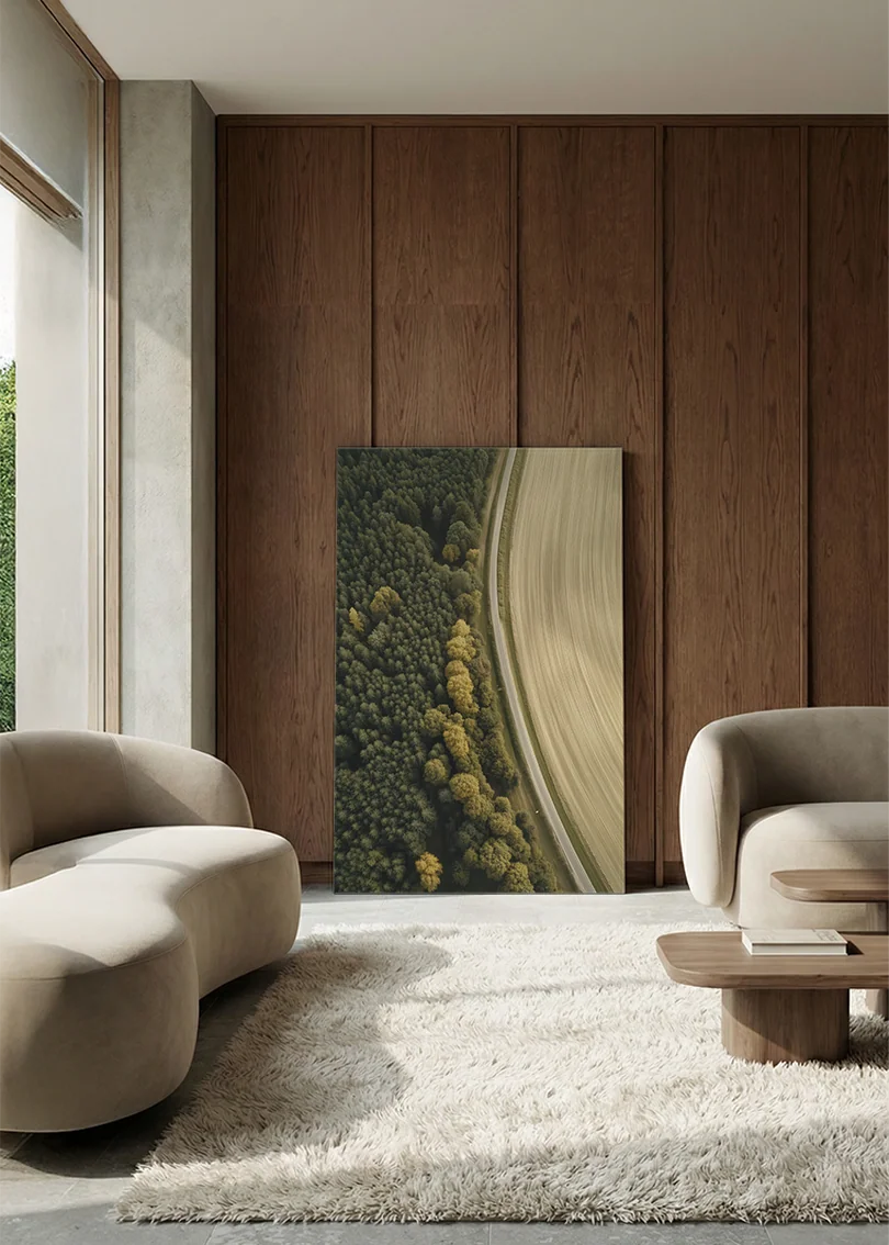 Akustikbillede med dronefoto af en buet markkant mellem skov og lys mark, i lounge med bugtet sofa mod vægpanel i valnød